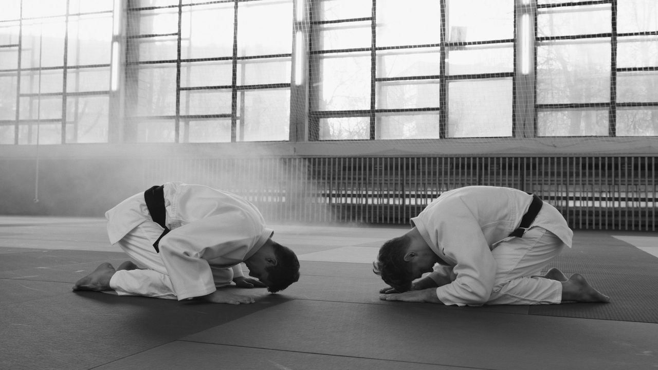 Two judoka in white gis and black belts kneel face to face in a seated bow, a tall wall of windows behind them