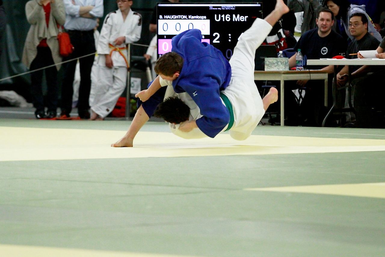 An under-16 judoka in a blue gi throws his opponent in white over his back